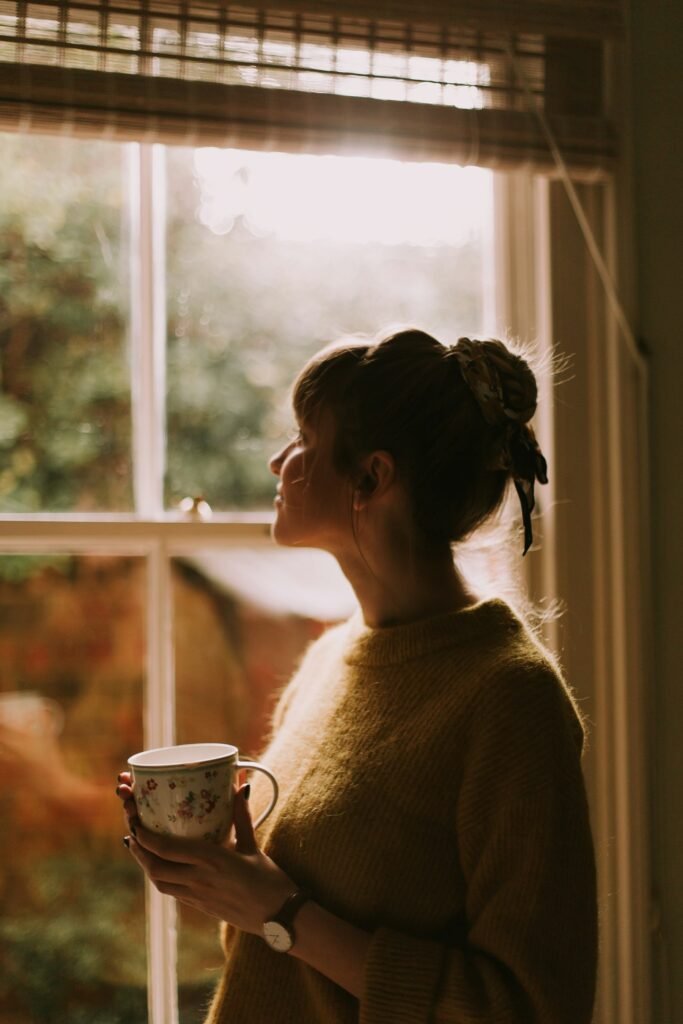 Mujer junto a una ventana con luz cálida sosteniendo una taza